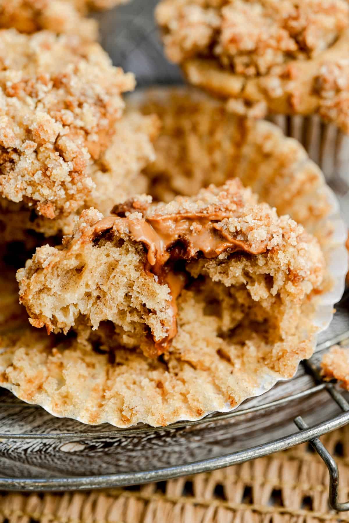 A close-up of a crumbly muffin broken in half, revealing a gooey caramel or peanut butter filling, sitting in a paper muffin liner on a rustic metal tray.