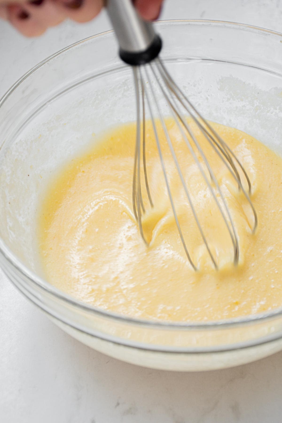 A hand holds a whisk, mixing a yellow batter in a clear glass bowl on a white surface.