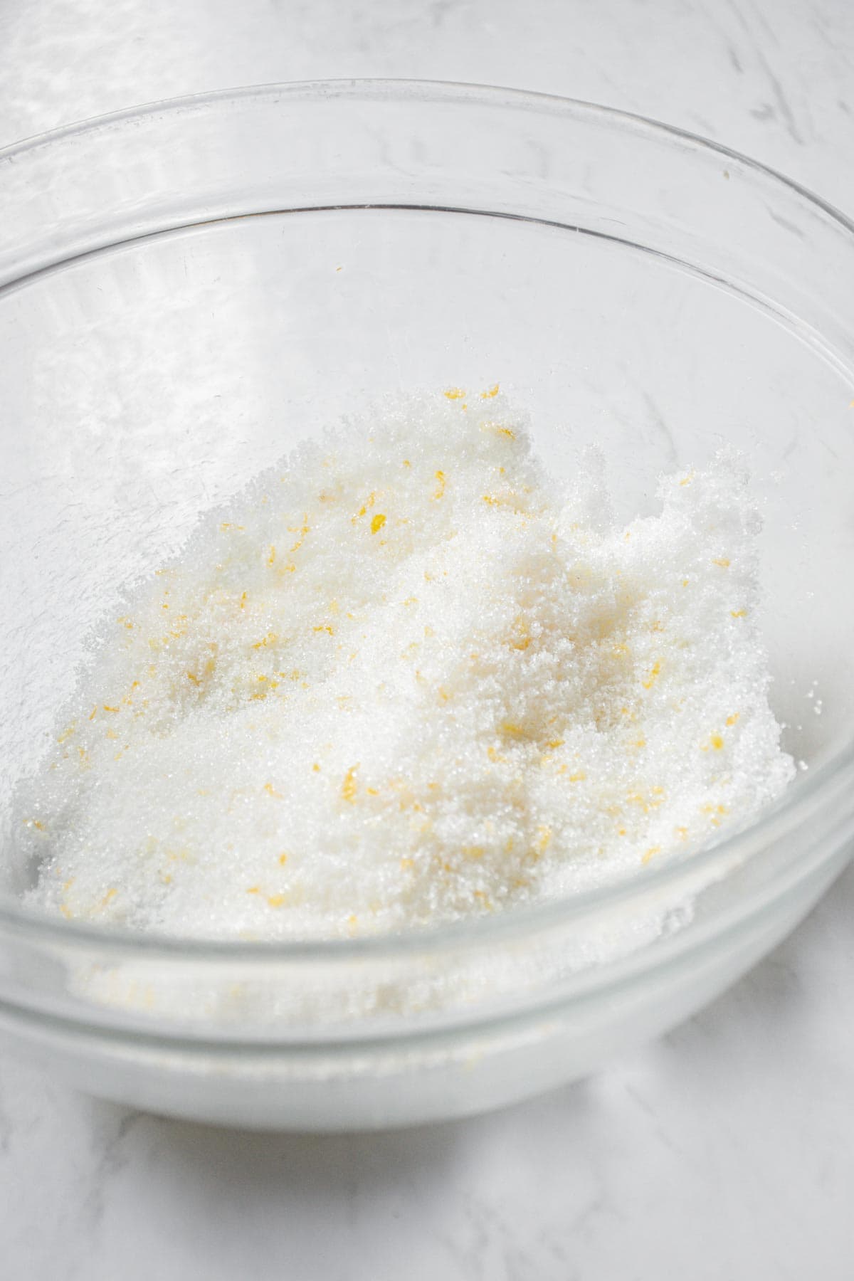 A clear glass bowl containing a mixture of white granulated sugar and small yellow lemon zest pieces, placed on a white marble surface.