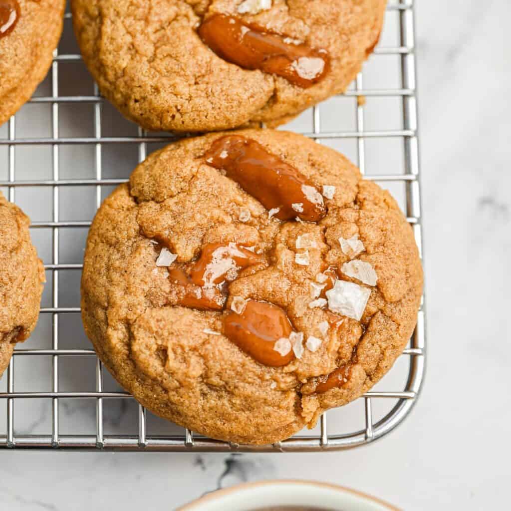 Close-up of caramel cookies topped with flaky sea salt resting on a metal cooling rack, with part of a white surface visible underneath.
