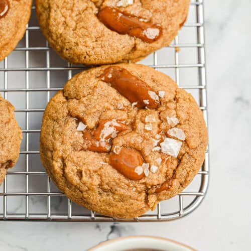 Close-up of caramel cookies topped with flaky sea salt resting on a metal cooling rack, with part of a white surface visible underneath.