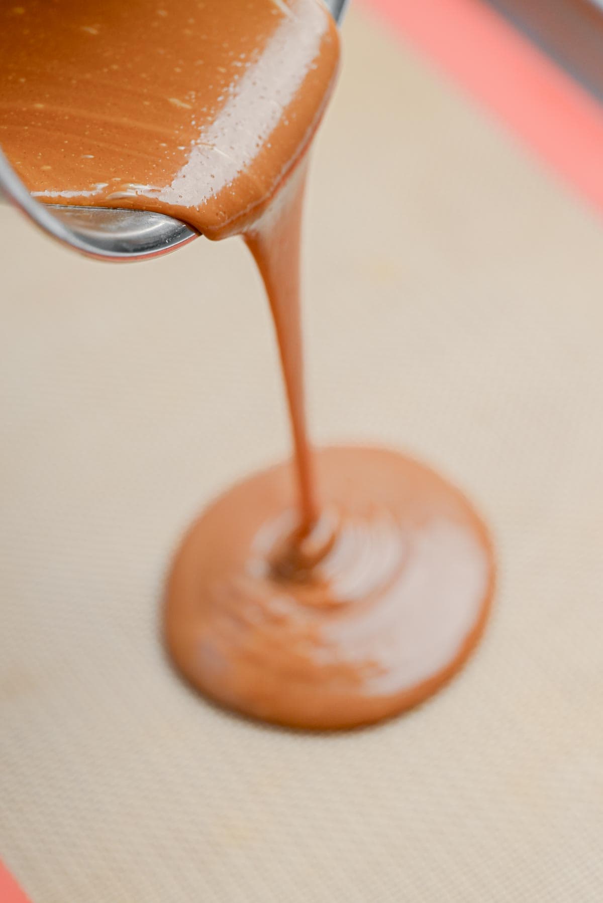 Thick caramel sauce, flecked with homemade toffee bits, is poured from a metal bowl onto a silicone baking mat, forming a smooth, glossy pool.
