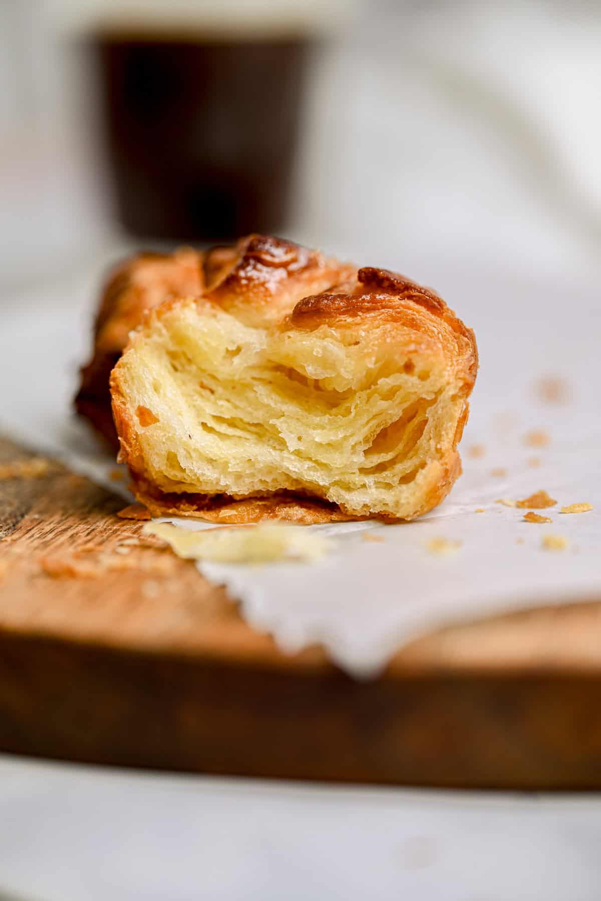 A close-up of a flaky, golden pastry with visible layers, resting on parchment paper atop a wooden board, with crumbs scattered around and a blurred cup in the background.