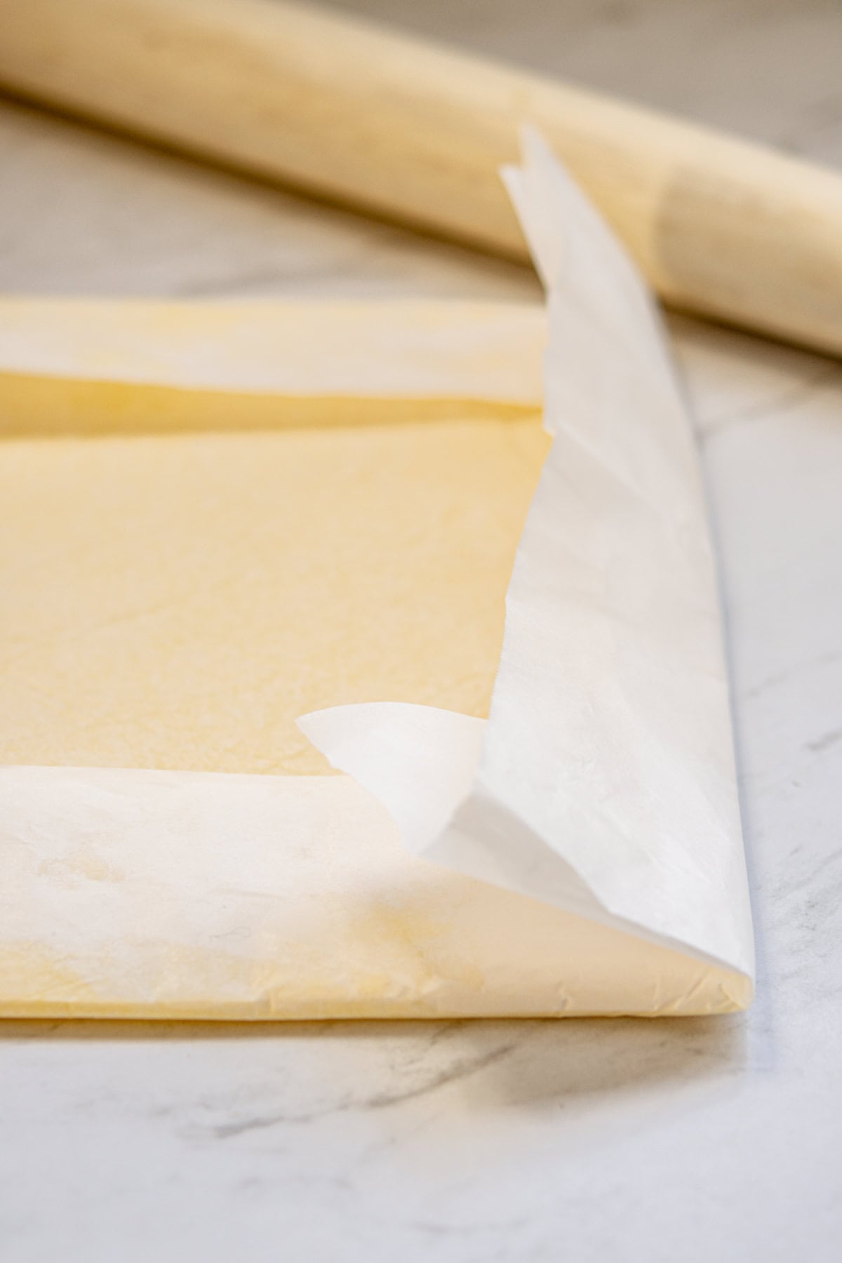 A sheet of dough lies on parchment paper, with one corner of the paper peeled back, and a wooden rolling pin is visible in the background on a marble countertop.