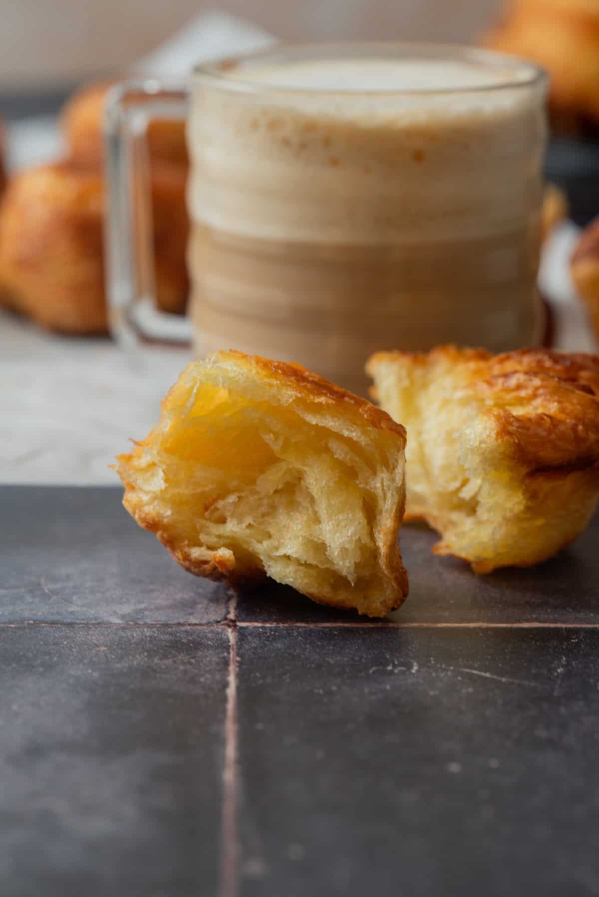 A close-up of a flaky, golden Kouign Amann broken in half sits on a dark surface, with a glass mug of frothy coffee blurred in the background.