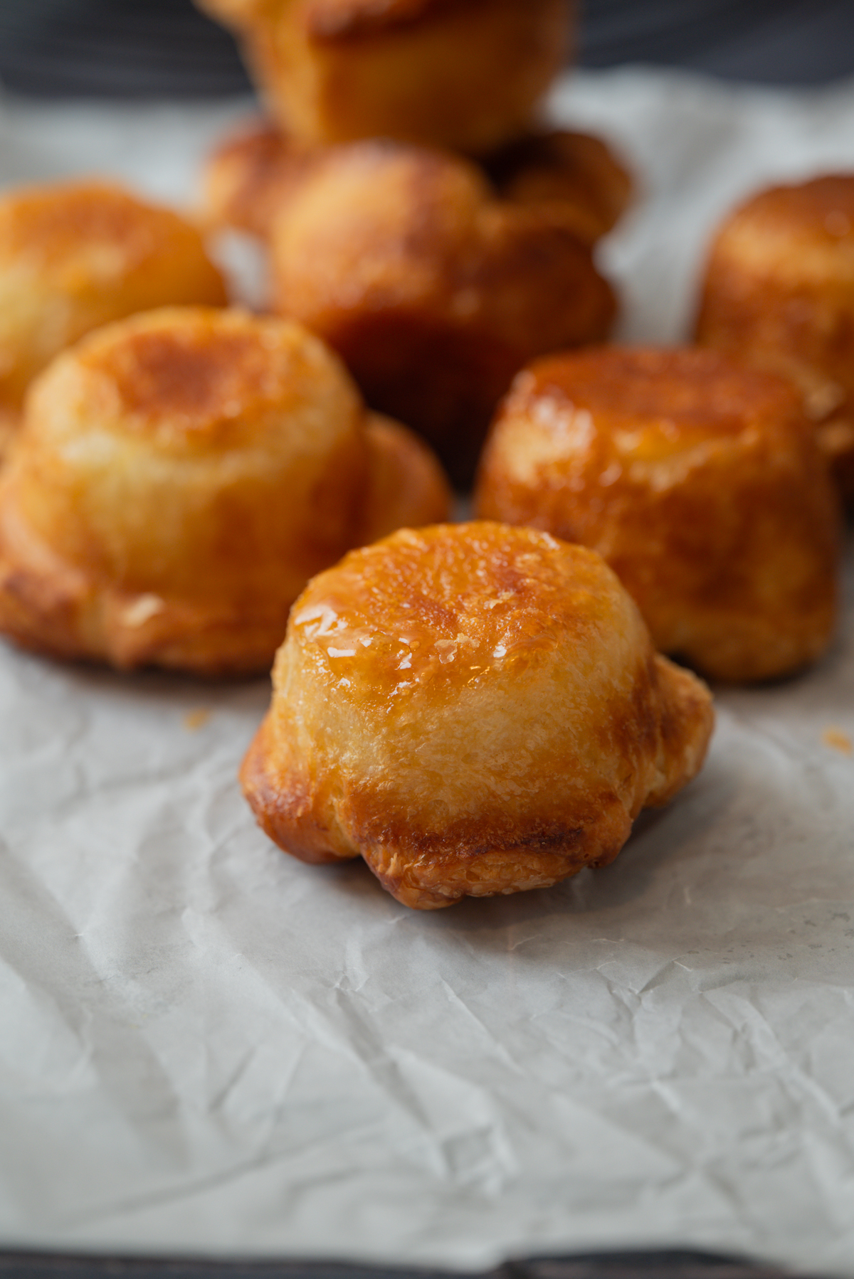 Golden-brown Kouign Amann pastries with a glossy finish are arranged on white parchment paper. These treats have a round, slightly domed shape and crisp, caramelized edges.