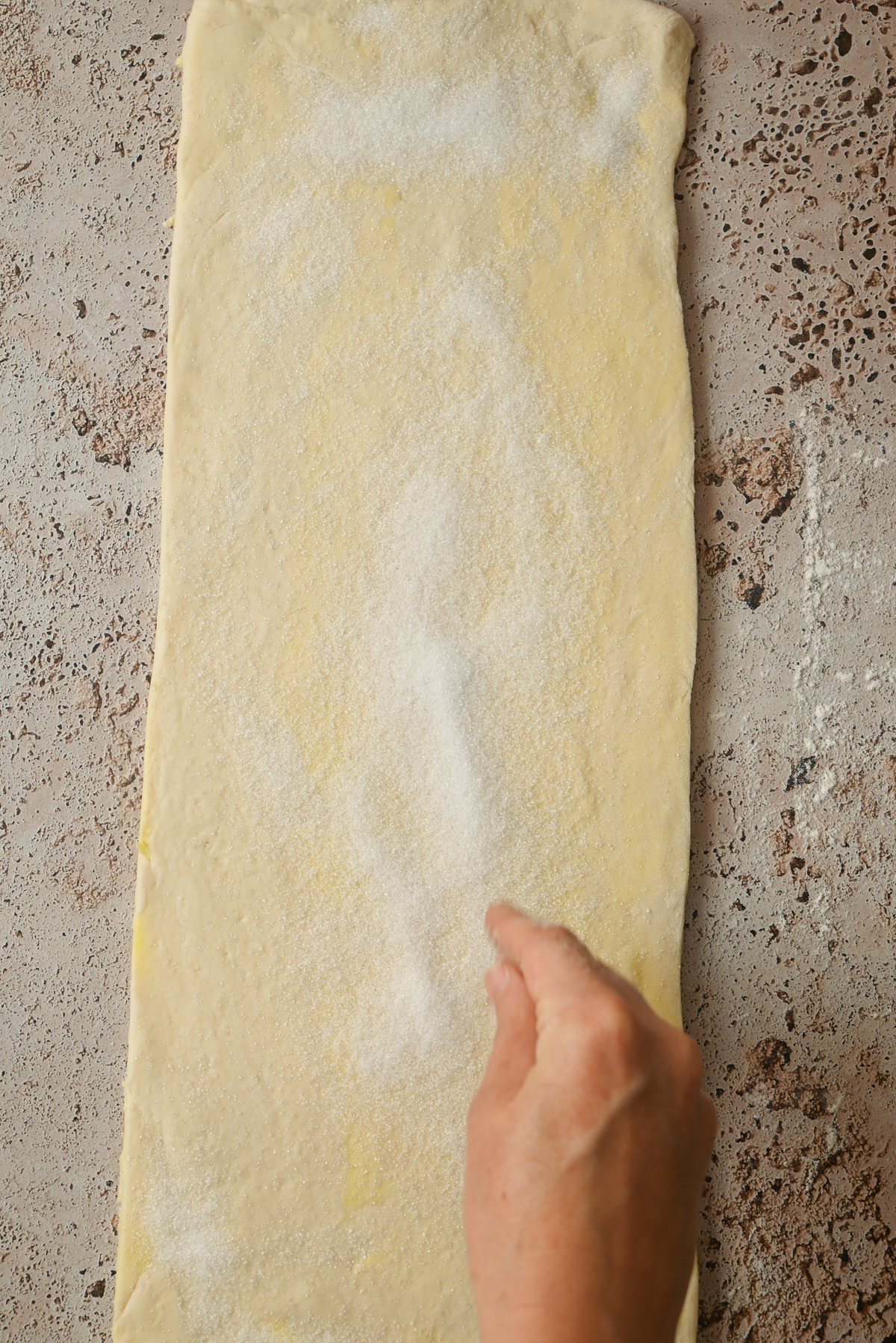 A hand sprinkles sugar over a large, rectangular sheet of rolled-out dough on a lightly floured, textured surface, preparing the base for Kouign Amann.