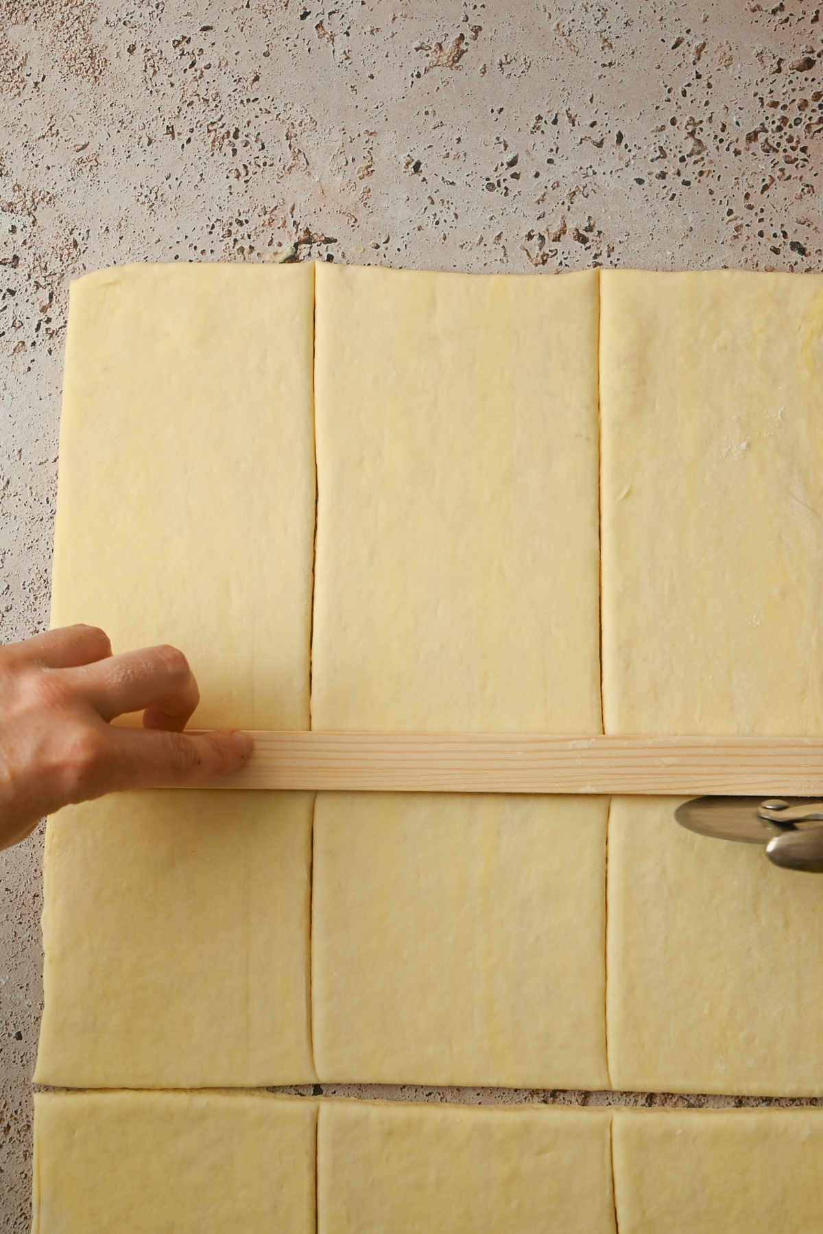 A hand holds a wooden ruler on top of a sheet of dough marked into rectangles for Kouign Amann, while a pastry cutter is used to cut along the ruler’s edge.