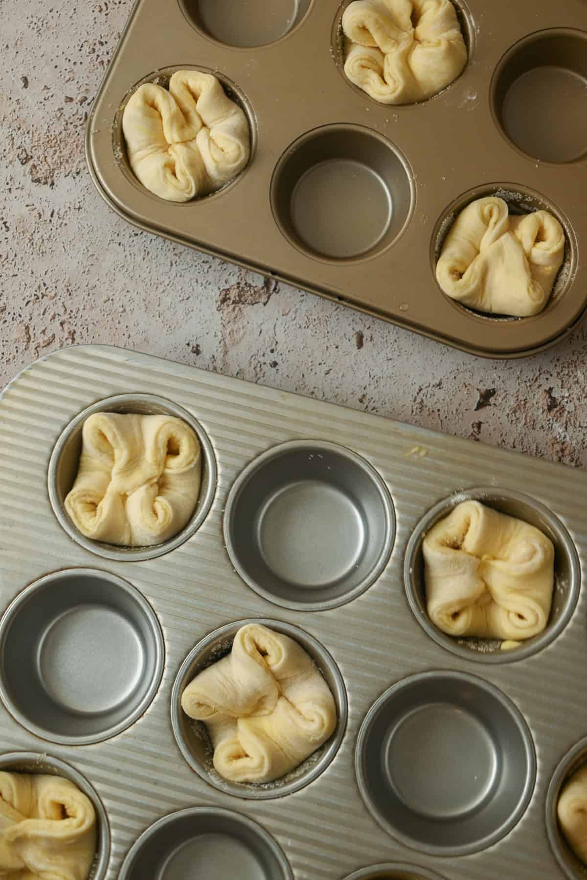 Two muffin pans, some cups filled with folded Kouign Amann dough and others empty, are displayed on a light, textured surface, ready to be baked to golden perfection.