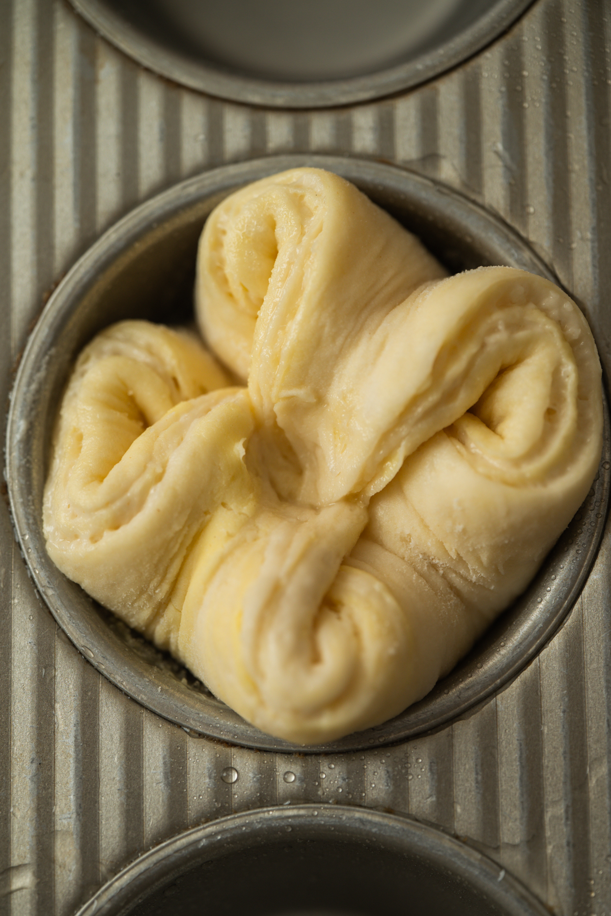 A close-up of a folded piece of raw Kouign Amann dough placed in a metal muffin tin, ready to be baked. The dough has a soft, slightly shiny surface and is arranged in a flower-like shape.