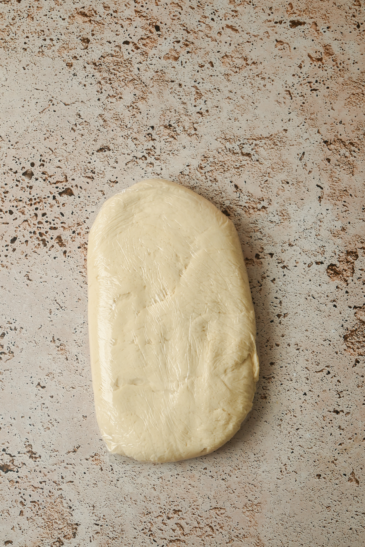 A rectangular portion of Kouign Amann dough wrapped in plastic wrap rests on a textured, light brown countertop.