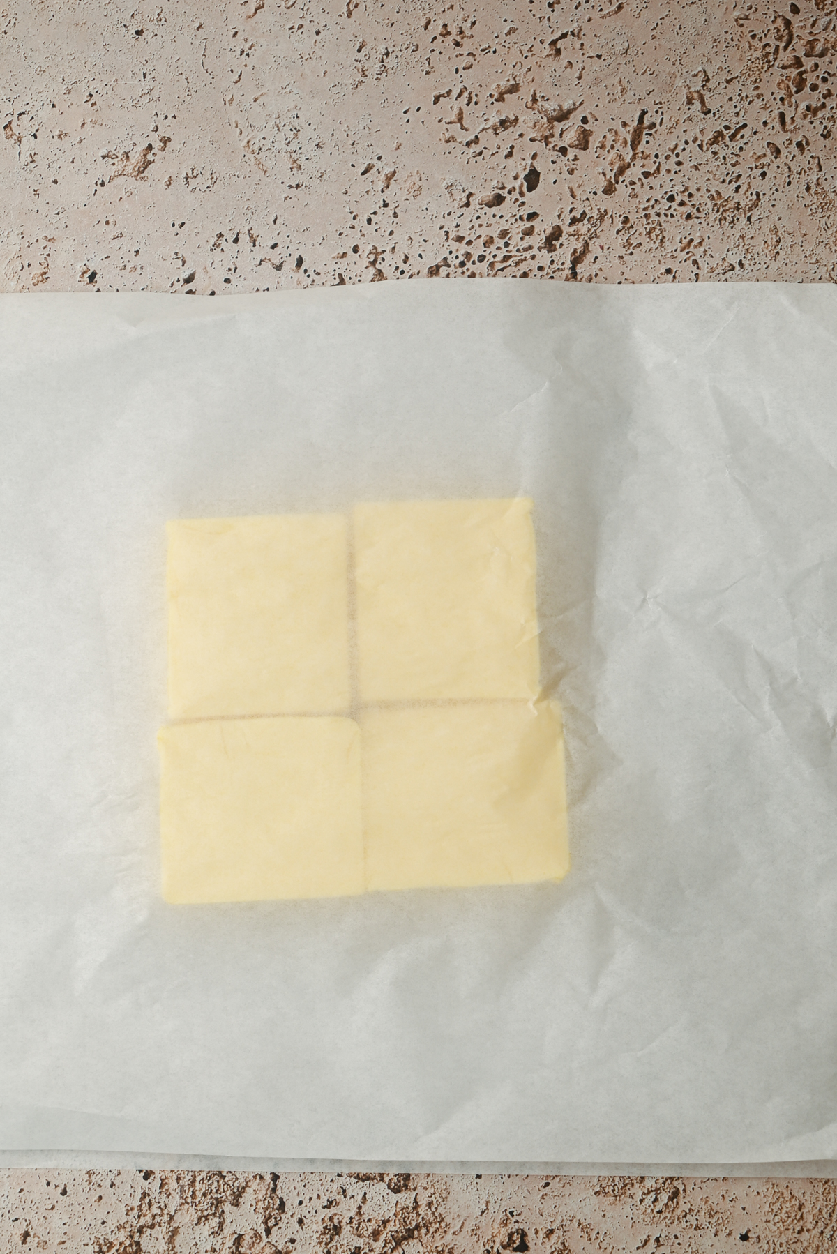 Four squares of butter placed together on a sheet of parchment paper, ready to create the rich layers found in kouign amann, on a textured beige surface.