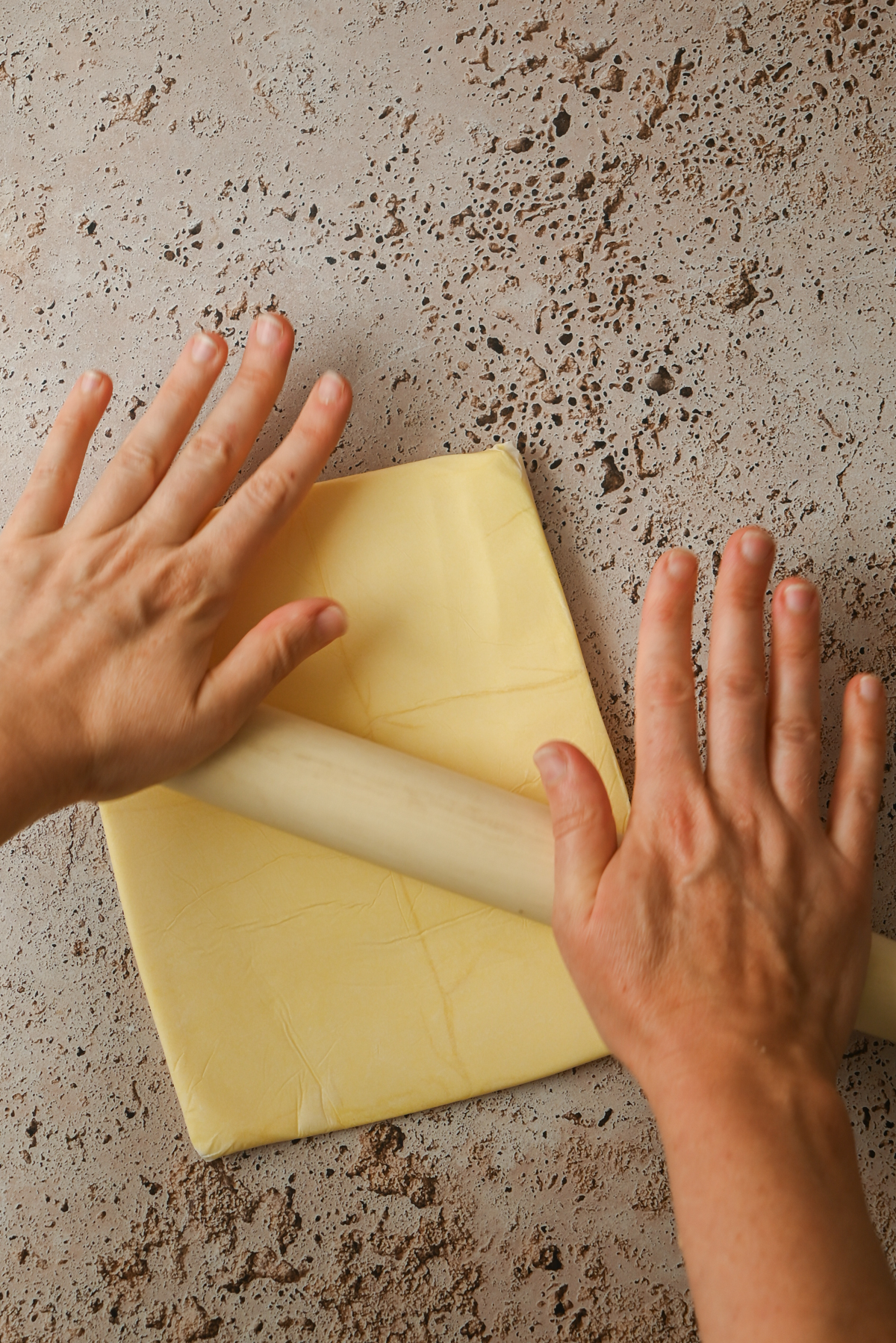 Two hands use a rolling pin to flatten Kouign Amann dough into a square on a speckled countertop.