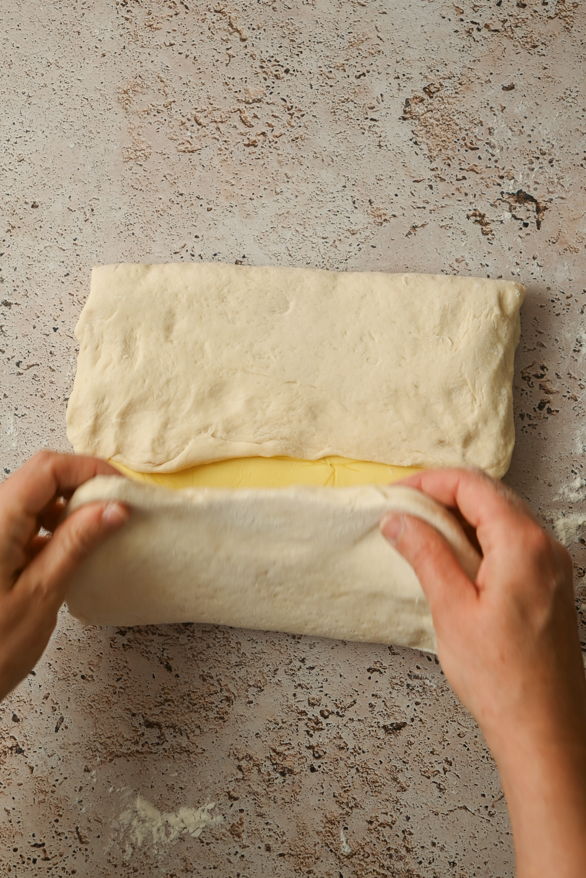 Hands folding a sheet of dough over butter on a textured countertop, preparing the base for Kouign Amann lamination.
