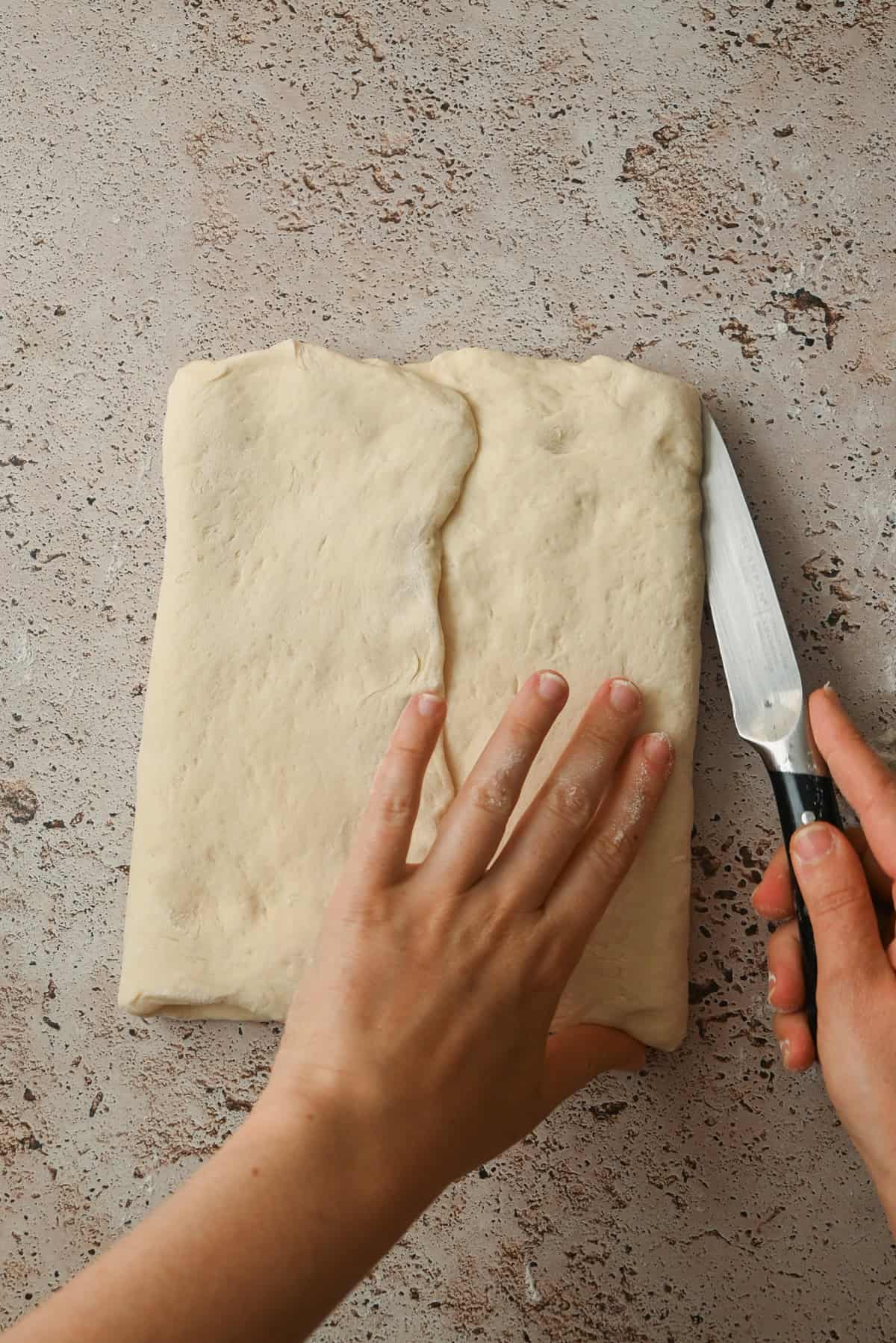 Two hands preparing dough for Kouign Amann, with one hand folding the dough and the other poised with a knife, ready to cut on a textured countertop.