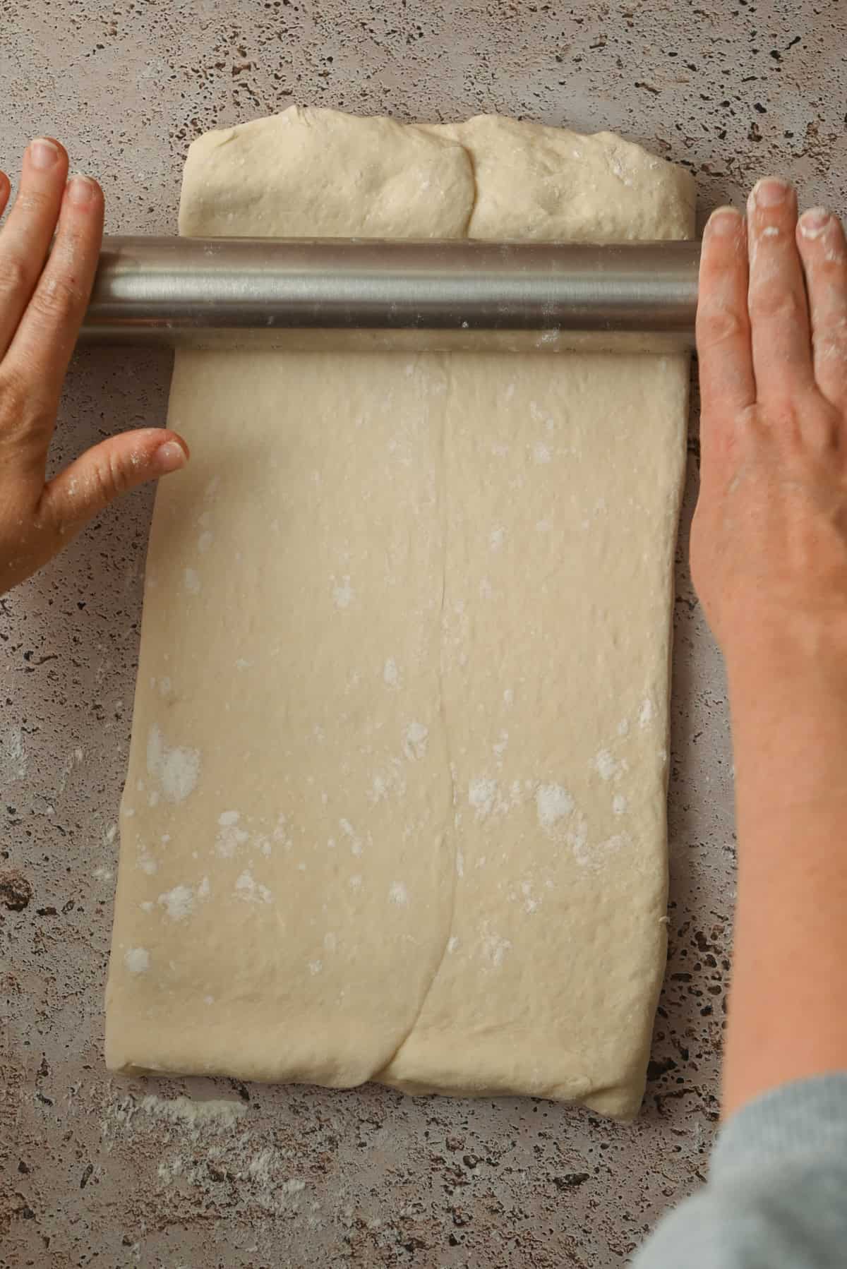 Two hands use a rolling pin to roll out a rectangle of dough for Kouign Amann on a floured surface.