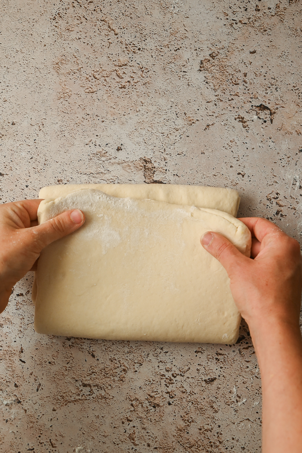 Two hands folding a sheet of uncooked, lightly floured dough on a textured, light-colored surface, preparing it for Kouign Amann.