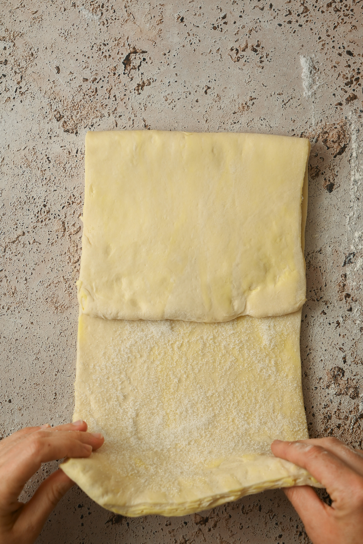 Two hands fold a sheet of dough on a lightly floured, textured surface, preparing buttery layers for pastry, croissants, or Kouign Amann.