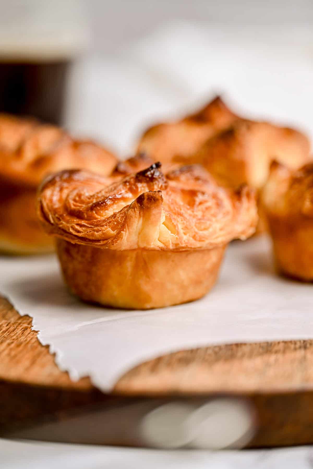 A close-up of golden, flaky, baked pastries with crispy tops, resting on a piece of parchment paper atop a wooden surface. The background is softly blurred, highlighting the pastry texture.
