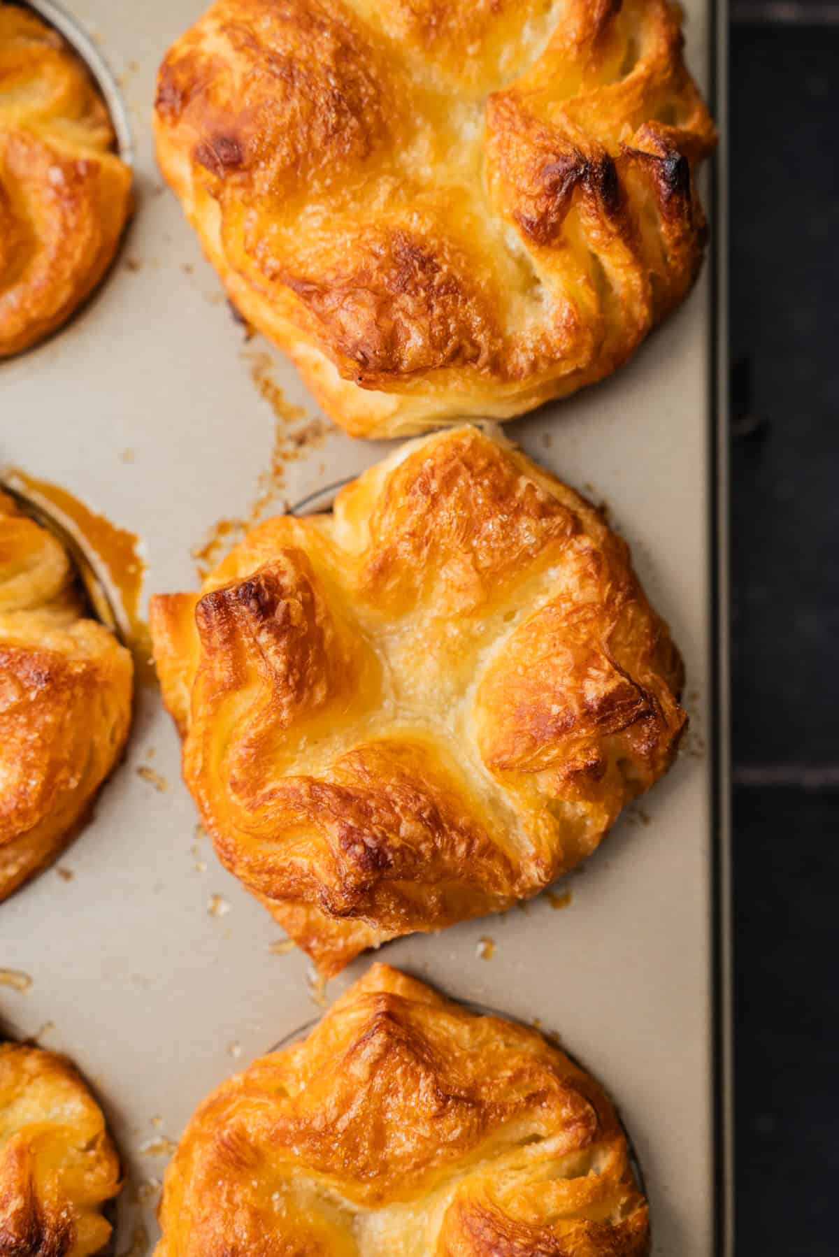 Golden, flaky Kouign Amann pastries with crisp tops in a muffin tin, fresh from the oven. The pastries are puffed and slightly browned, with visible buttery layers and some caramelized edges.