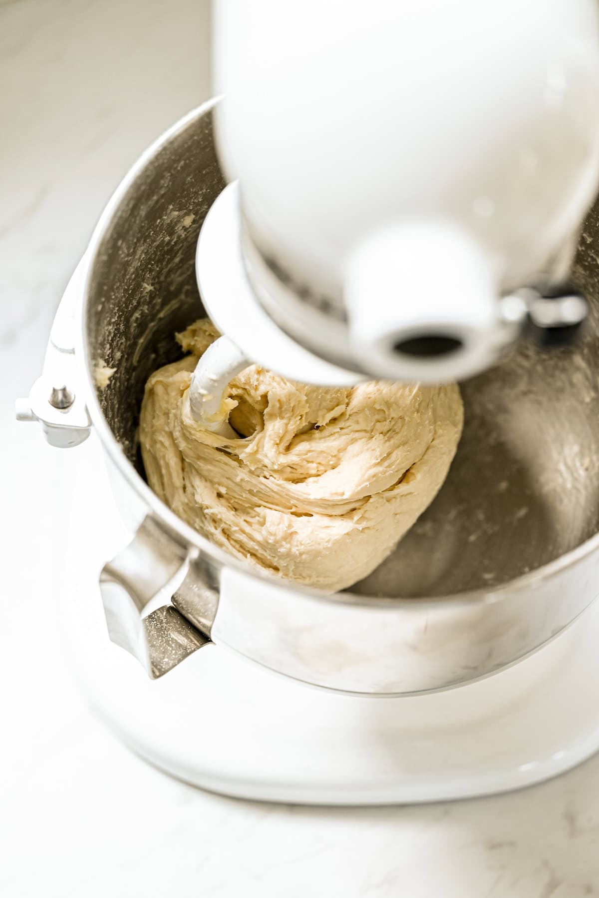 A stand mixer with a dough hook attachment is mixing bread dough in a stainless steel bowl on a white countertop.