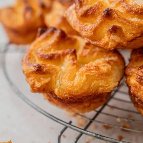 Close-up of golden, flaky Kouign Amann pastries with crispy edges stacked on a wire cooling rack, with crumbs scattered on the light surface below.