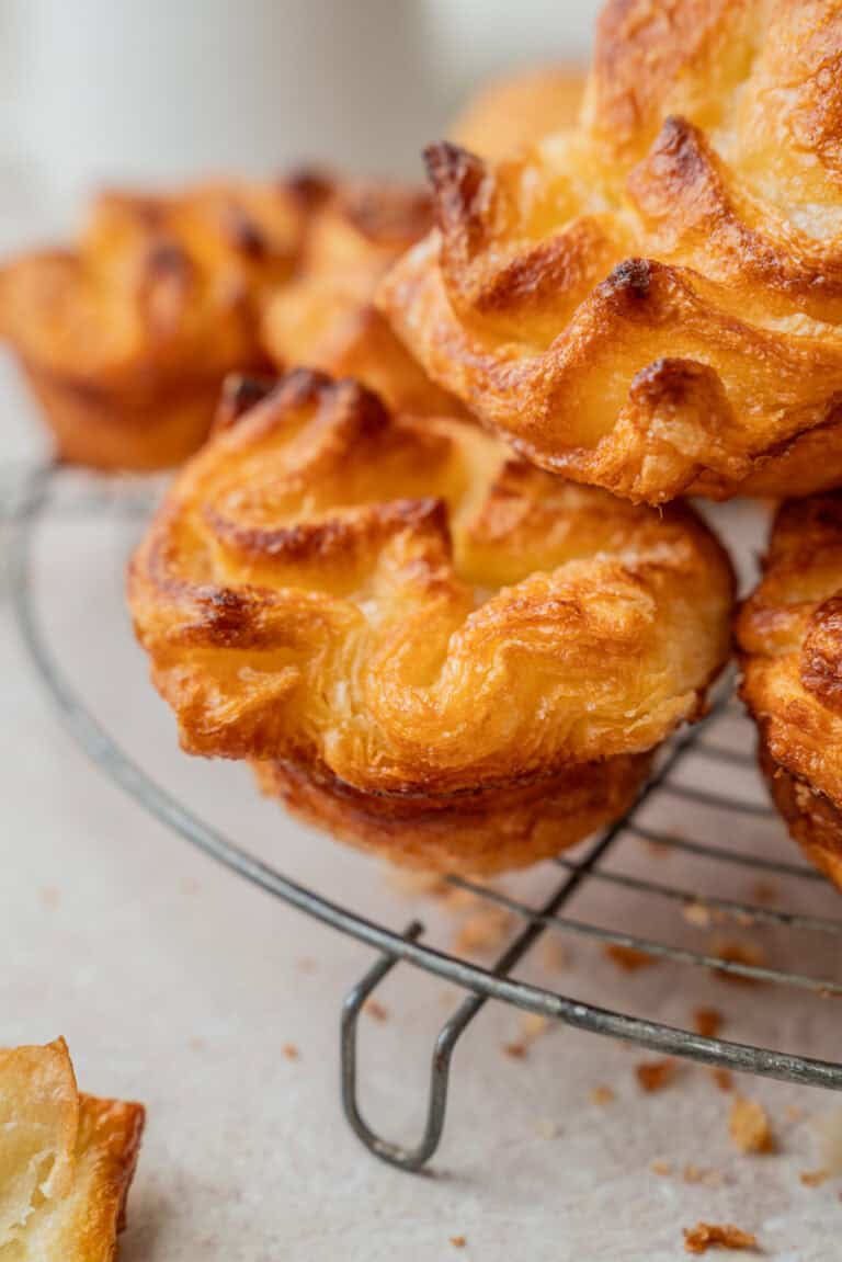 Close-up of golden, flaky Kouign Amann pastries with crispy edges stacked on a wire cooling rack, with crumbs scattered on the light surface below.