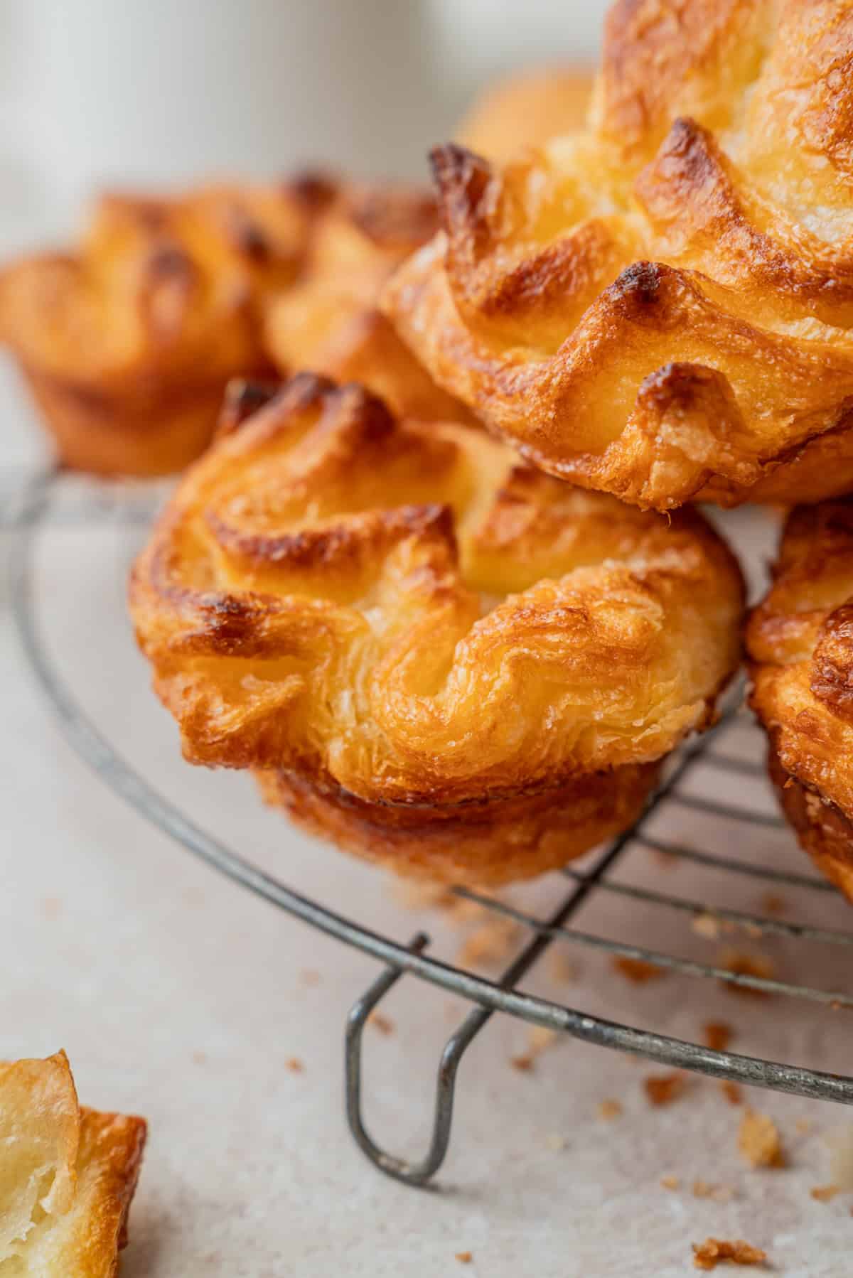 Close-up of golden, flaky Kouign Amann pastries with crispy edges stacked on a wire cooling rack, with crumbs scattered on the light surface below.