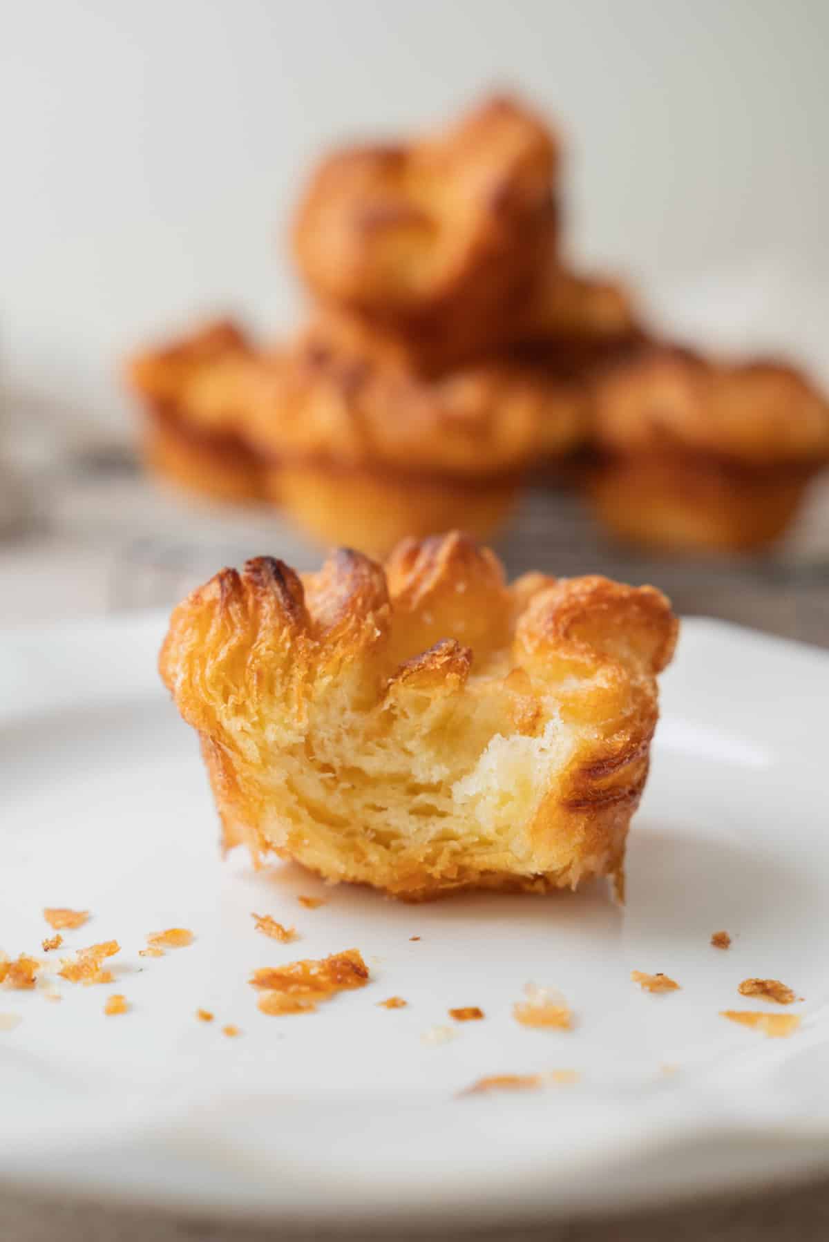 A golden, flaky Kouign Amann pastry with a bite taken out sits on a white plate, with a few crumbs scattered around. More pastries are blurred in the background.