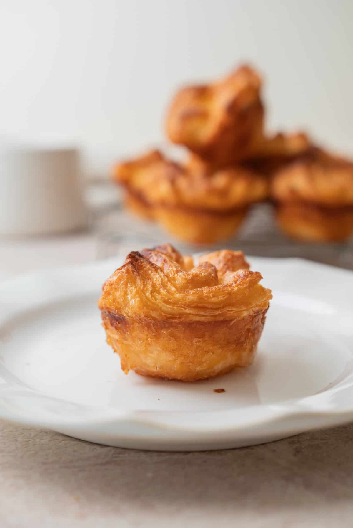 A golden, flaky Kouign Amann pastry sits on a white plate in the foreground, with more pastries stacked on a wire rack in the blurred background.