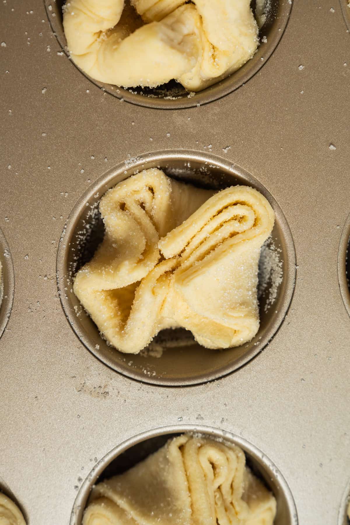 Unbaked Kouign Amann pastry dough folded into a delicate flower shape sits in a muffin tin cup, sprinkled lightly with sugar.
