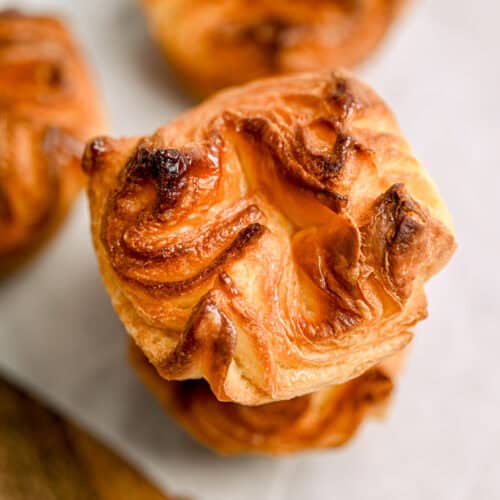 Close-up of two golden-brown, flaky pastries stacked on parchment paper, with another pastry blurred in the background. The pastries have a crispy, swirled surface and are set on a wooden surface.