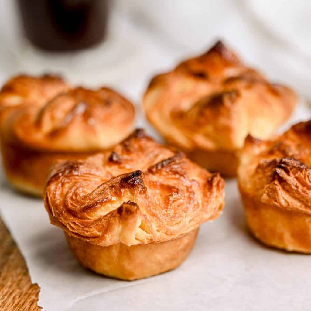 Four golden-brown, flaky pastries with a crimped top sit on parchment paper, showcasing their crisp, layered texture. One pastry is in the foreground, with the others slightly blurred in the background.