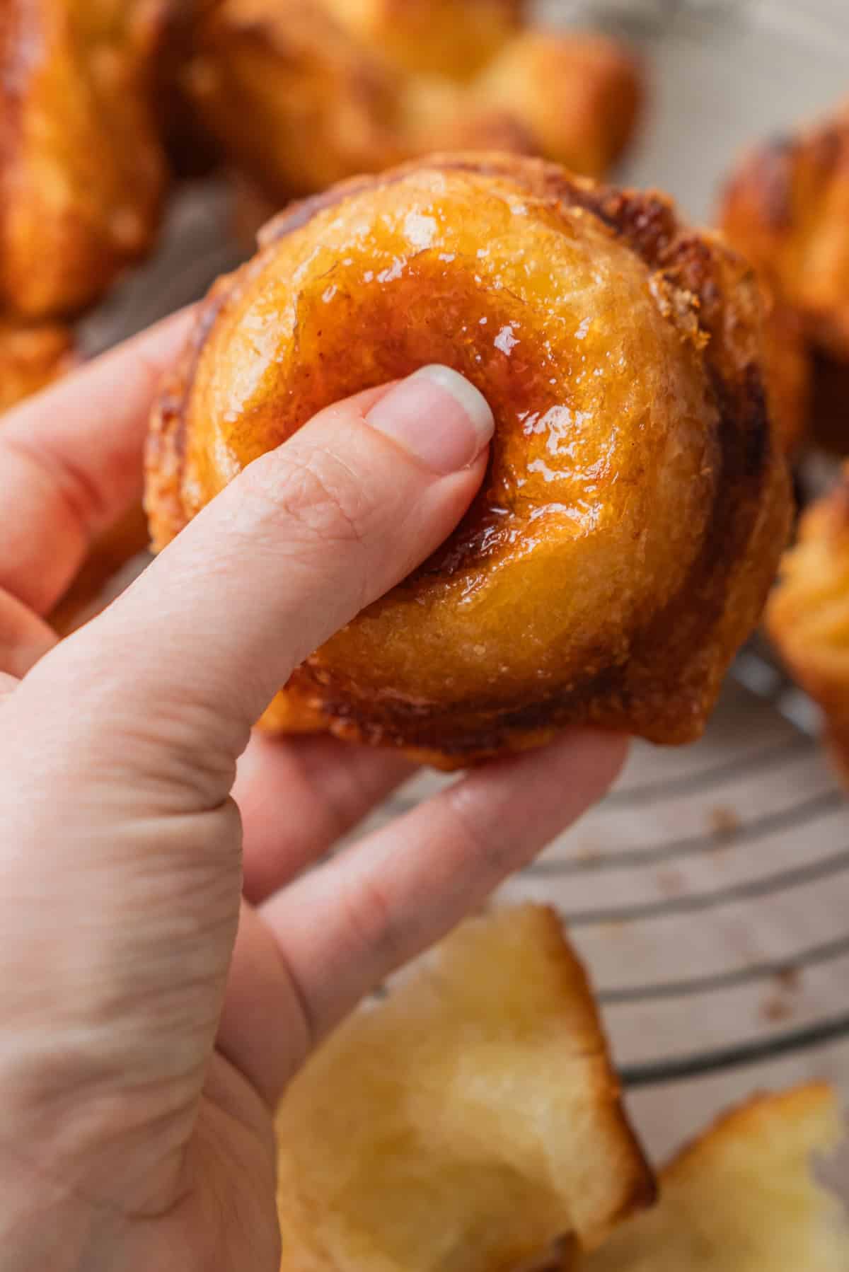 A hand holding a golden, glazed Kouign Amann pastry with a shiny surface, in front of a cooling rack with more Kouign Amann pastries in the background.