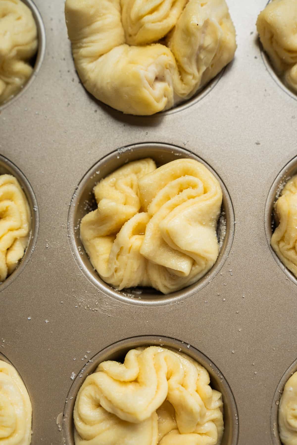 Unbaked folded Kouign Amann dough pieces are placed in a metal muffin tin, ready to be baked. The dough looks soft and slightly dusted with flour or sugar.
