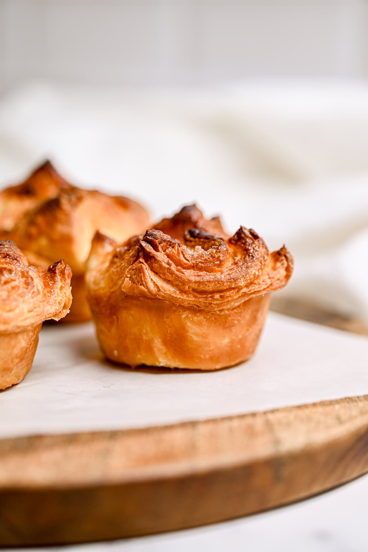 Golden brown, flaky pastries with crisp, textured tops are displayed on a round wooden board with a light, neutral background.