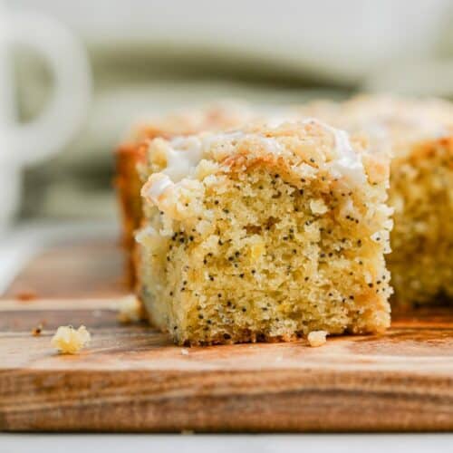 A close-up of a moist lemon poppy seed coffee cake square with a light icing glaze, resting on a wooden cutting board. The cake has a fluffy texture and visible poppy seeds throughout.