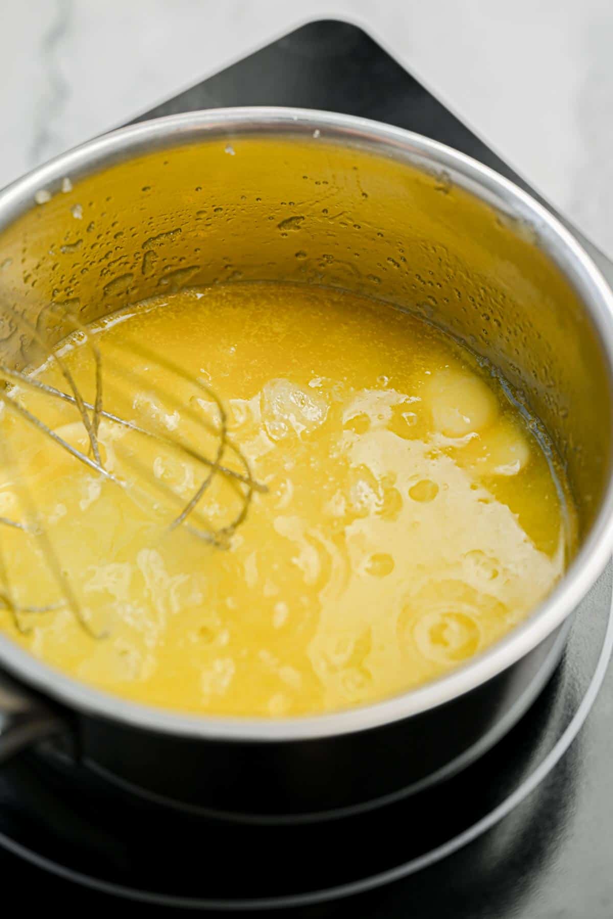 A close-up of a saucepan filled with a yellow, melted mixture being whisked on a stovetop.