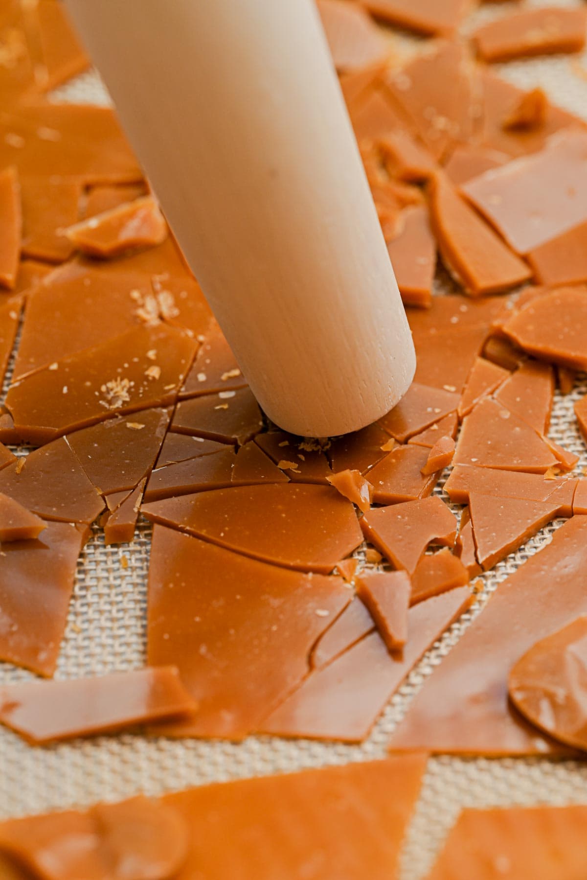 A close-up of a rolling pin crushing homemade toffee bits on a textured baking mat.