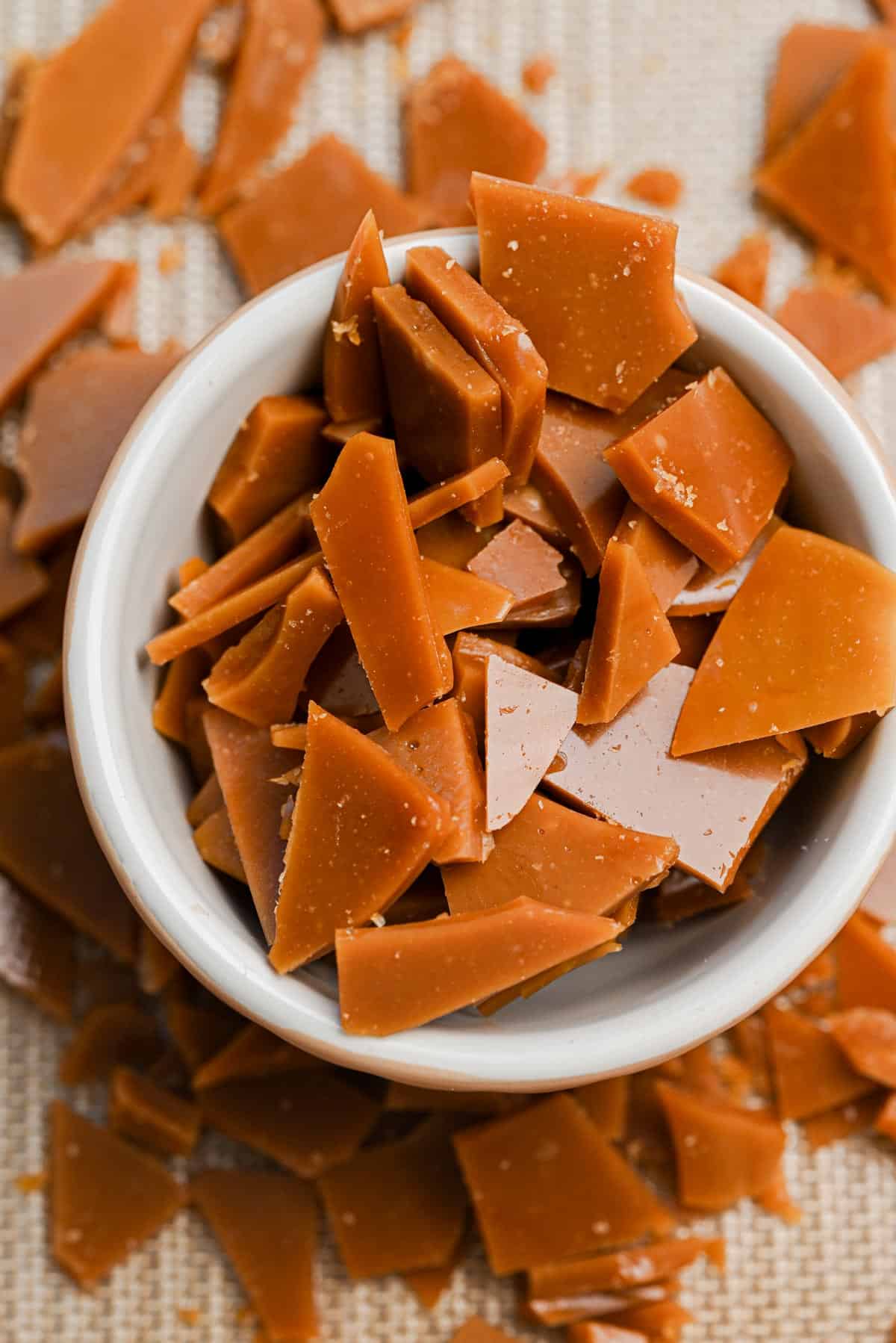 A white bowl filled with irregular shards of golden-brown homemade toffee bits, with more toffee pieces scattered on a textured surface around the bowl.