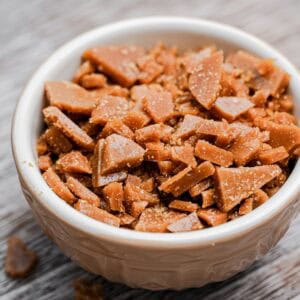 A white bowl filled with homemade toffee bits sits on a wooden surface, with some small pieces scattered nearby. The toffee is golden brown and has a crumbly, textured appearance.