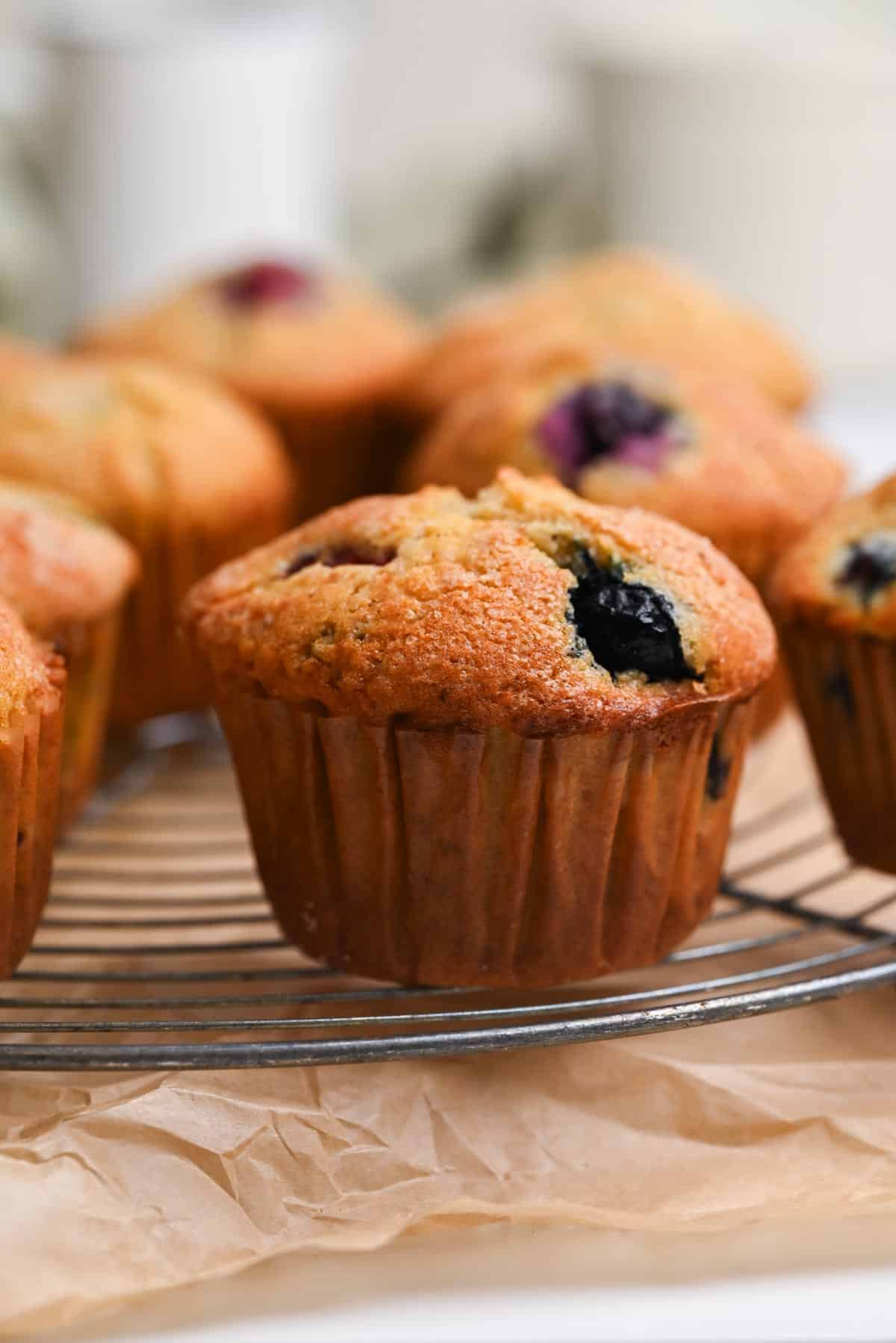 Close-up of freshly baked frozen berry muffin in brown paper liners cooling on a wire rack, with a few more muffins blurred in the background.