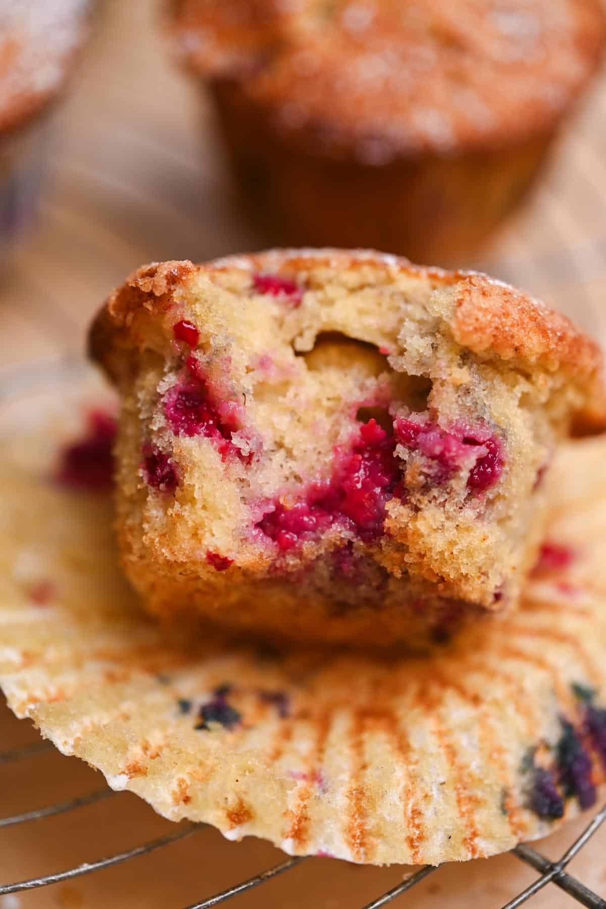 A close-up of a frozen berry muffin with visible raspberries inside, cut in half and resting on an opened muffin wrapper atop a metal cooling rack. The muffin looks moist, with a golden-brown top and vibrant red fruit pieces.