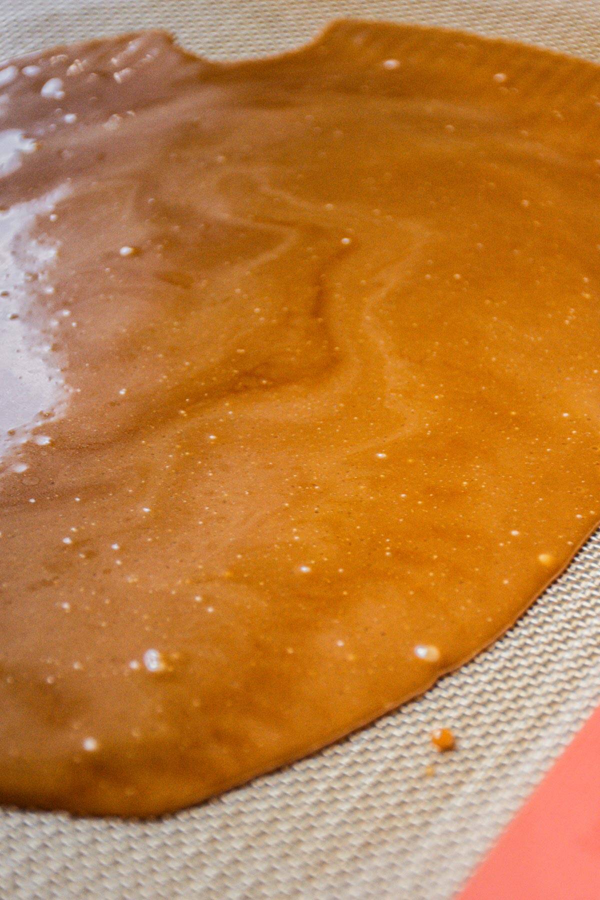 A sheet of caramel-colored homemade toffee bits spread thinly on a silicone baking mat, with small bubbles and a slightly glossy surface visible.