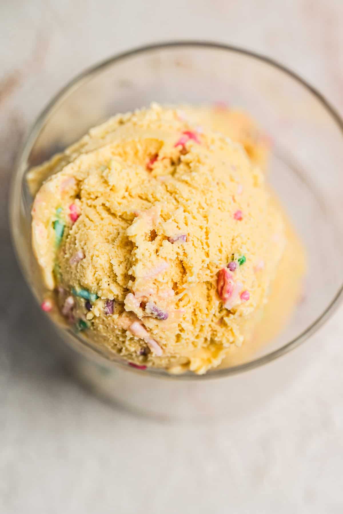 A close-up of a scoop of yellow ice cream with colorful sprinkles in a glass bowl on a light surface.