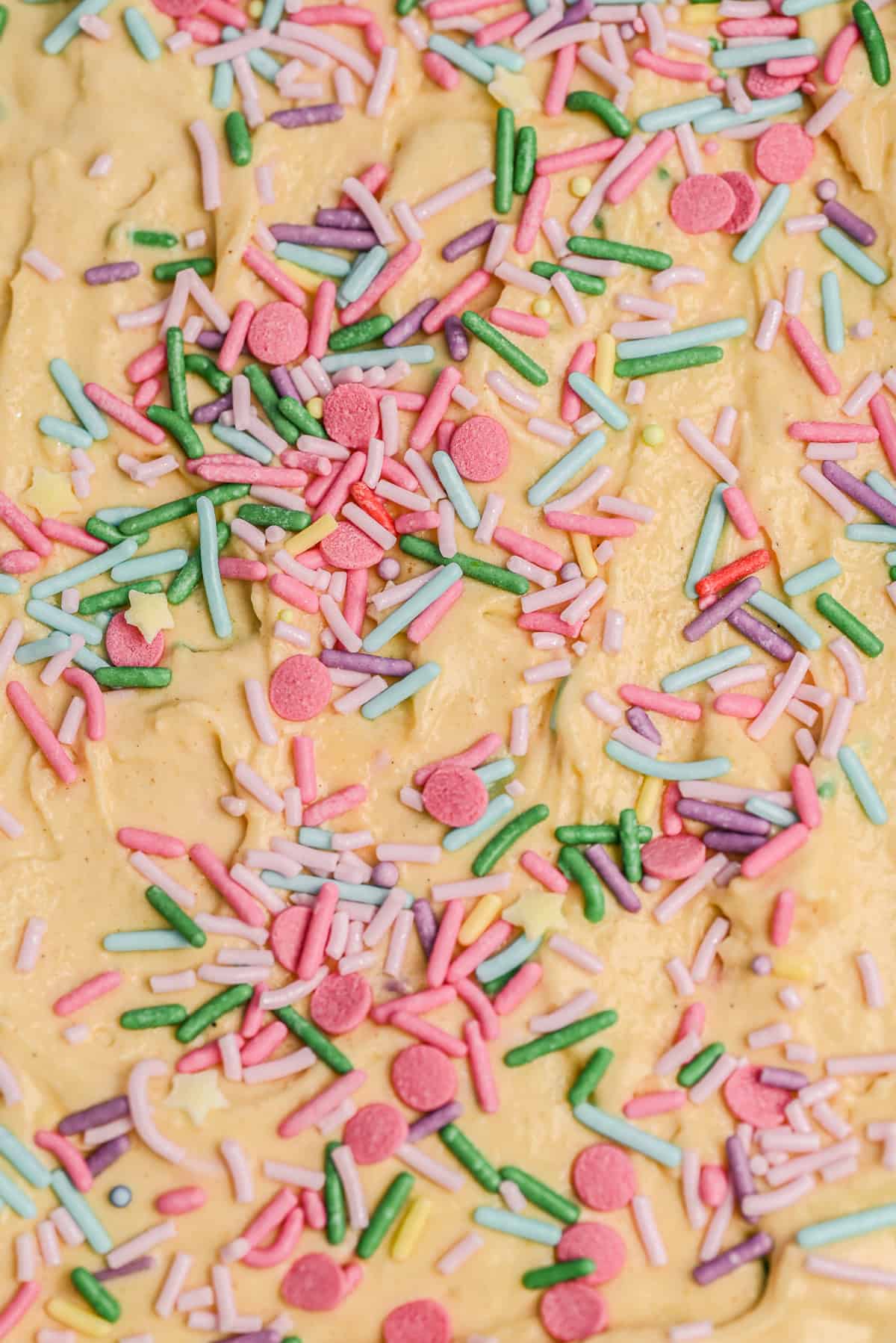 Close-up of yellow ice cream topped with colorful sprinkles, including pink, purple, green, and blue. The sprinkles are scattered unevenly across the surface.
