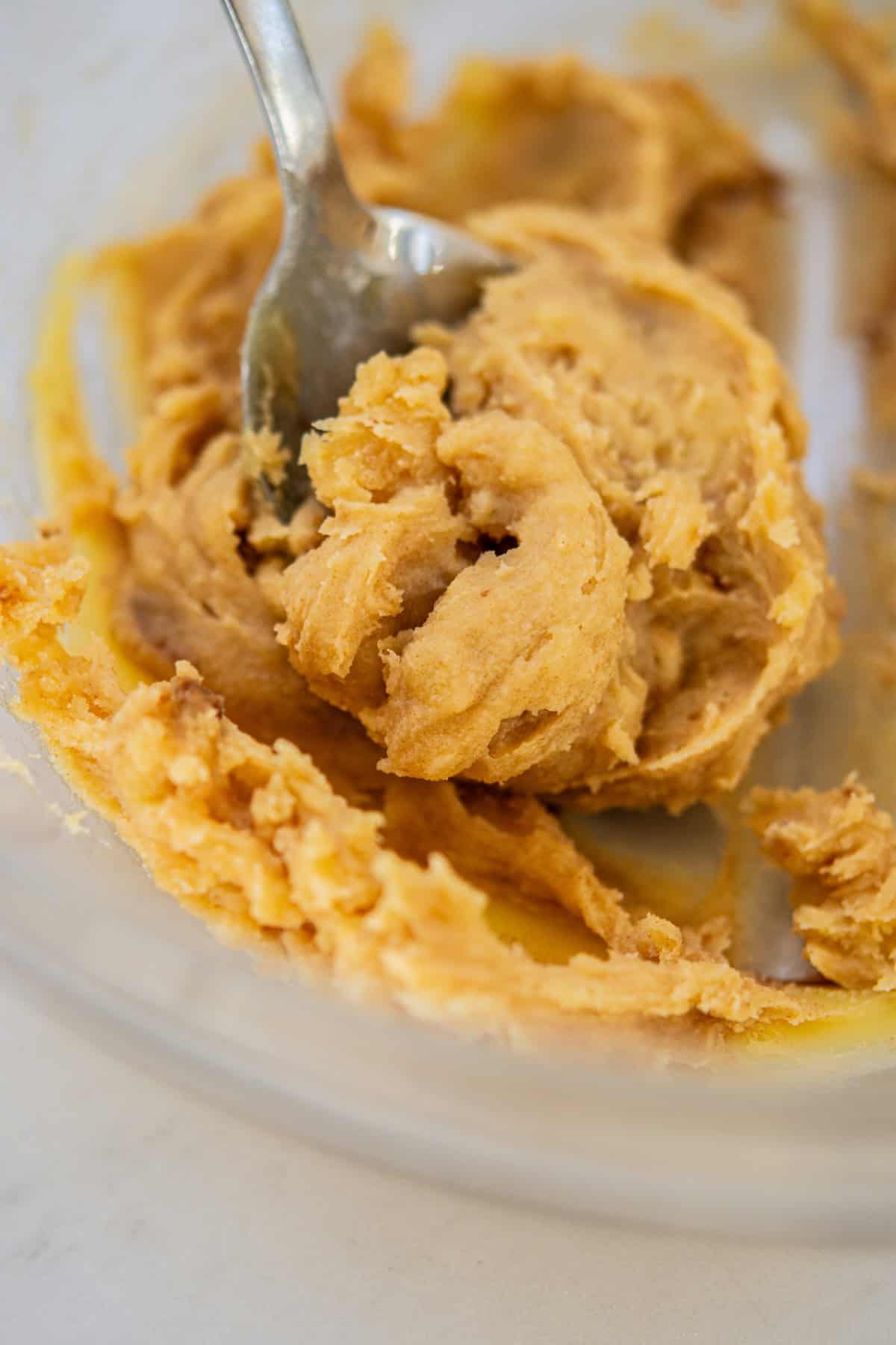 Close-up of a metal spoon mixing thick, creamy peanut butter cookie dough in a clear bowl. The dough has a light brown color and a smooth, slightly textured appearance.