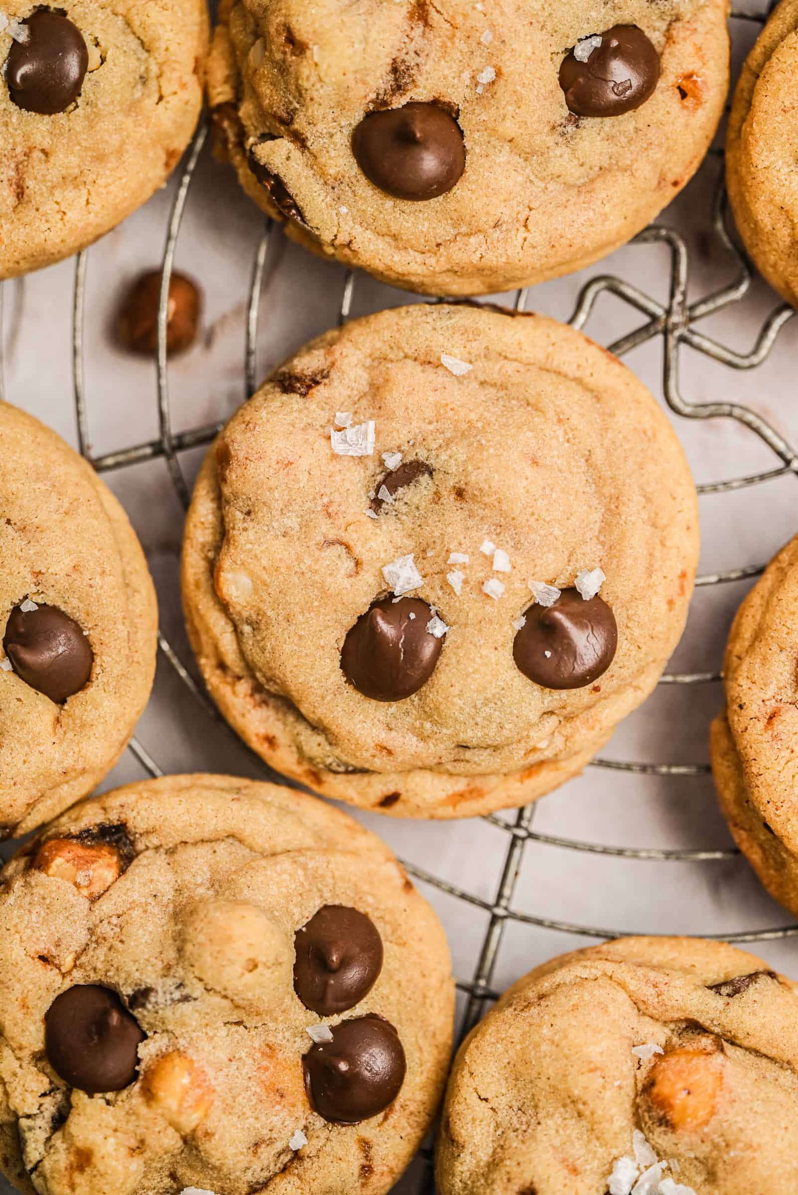A close-up of thick chocolate chip cookies on a wire cooling rack, sprinkled with flaky sea salt. The cookies have golden edges and visible chocolate chips.