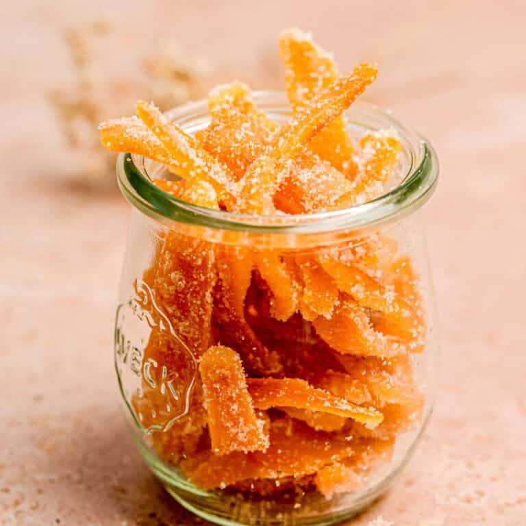 A glass jar filled with sugar-coated orange candied ginger strips sits on a light-colored surface, with a subtle, blurred background.