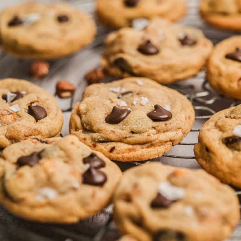 A close-up of freshly baked chocolate chip hazelnut cookies with flakes of sea salt on top, cooling on a wire rack. The cookies are golden brown and soft, with visible chocolate chips.